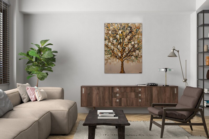 May include: A painting of a gold tree with a textured, abstract style. The tree is set against a light brown background. The painting is framed and hanging on a white wall in a living room setting.