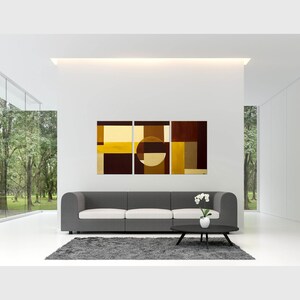 May include: Three abstract paintings in a modern living room. The paintings are in shades of brown, gold, and yellow. The paintings are hung on a white wall above a gray sofa. The sofa is in front of a large window with a view of trees.