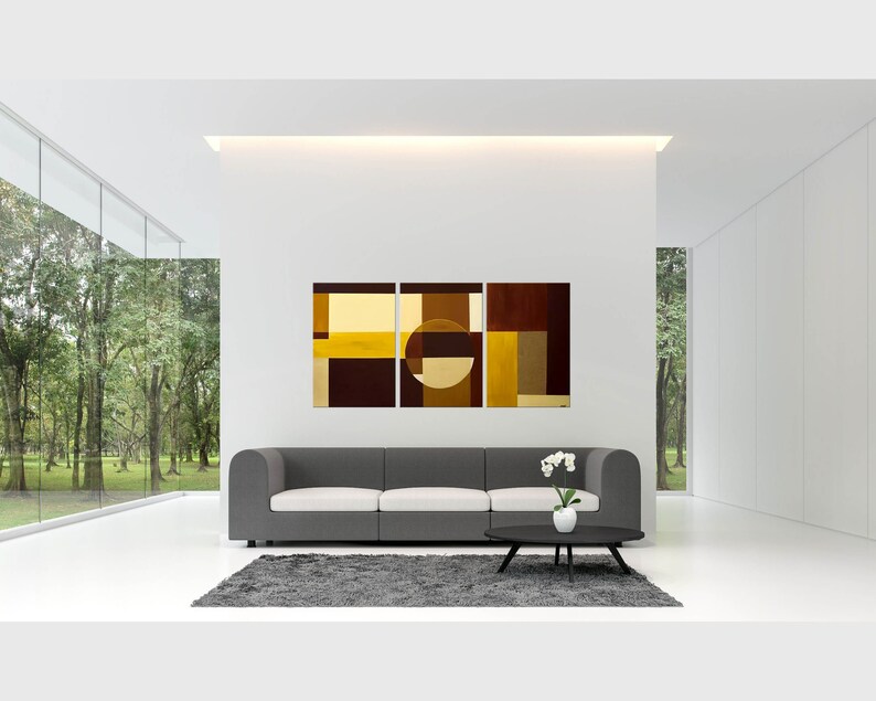 May include: Three abstract paintings in a modern living room. The paintings are in shades of brown, gold, and yellow. The paintings are hung on a white wall above a gray sofa. The sofa is in front of a large window with a view of trees.