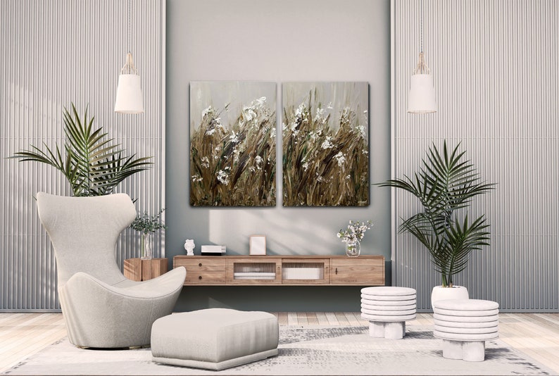 May include: A living room interior design with a white armchair and ottoman, two white side tables, a wooden media console, and two abstract paintings of white flowers on a brown background.