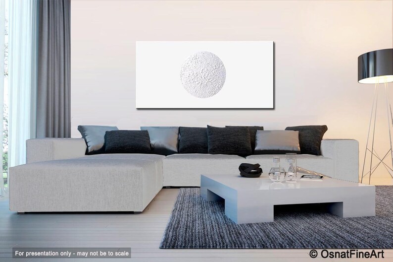 May include: White textured canvas painting with a large white circle in the center. The painting is hanging on a white wall above a gray sectional sofa with black pillows. A white coffee table sits in front of the sofa.