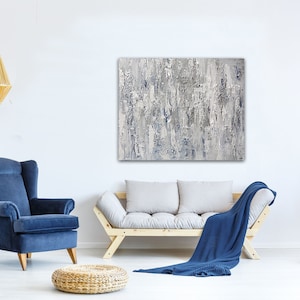 May include: A living room interior design with a blue armchair, a light gray sofa with a blue throw blanket, a woven ottoman, a hanging light fixture, and a large abstract painting in shades of gray and blue.