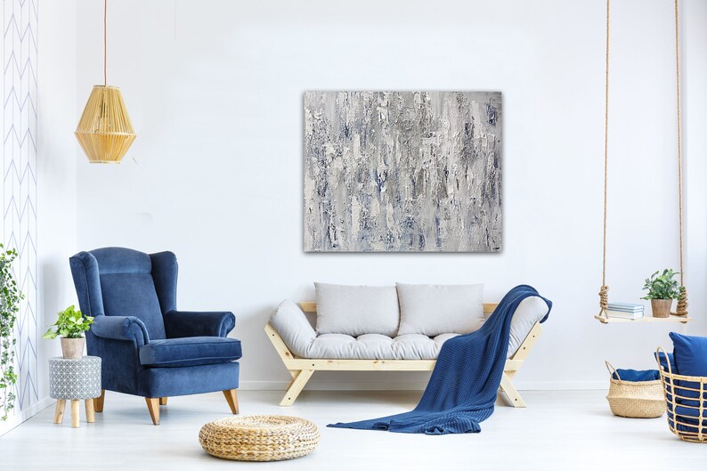 May include: A living room interior design with a blue armchair, a light gray sofa with a blue throw blanket, a woven ottoman, a hanging light fixture, and a large abstract painting in shades of gray and blue.