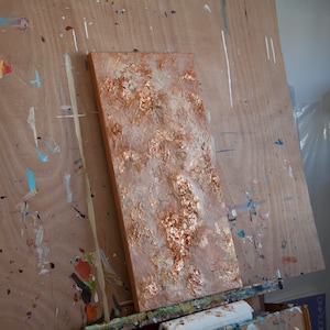 May include: A textured canvas painting with a gold and white abstract design. The painting is on an easel in an art studio.
