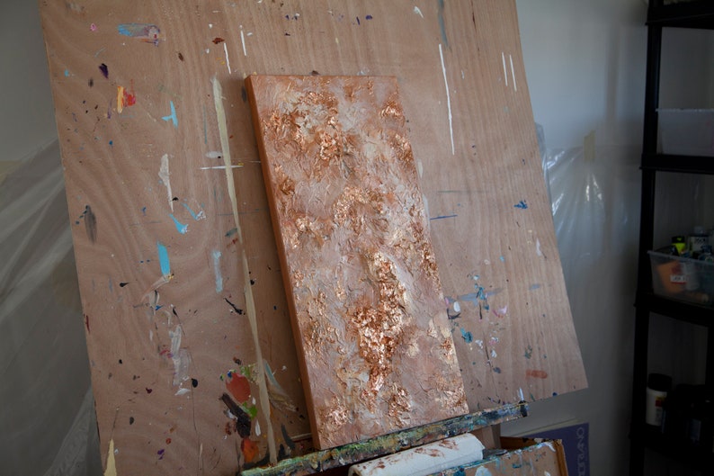 May include: A textured canvas painting with a gold and white abstract design. The painting is on an easel in an art studio.