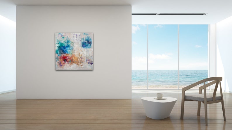 May include: Abstract painting with blue, red, orange, and yellow colors on a white background. The painting is hanging on a white wall in a modern room with a large window overlooking the ocean.