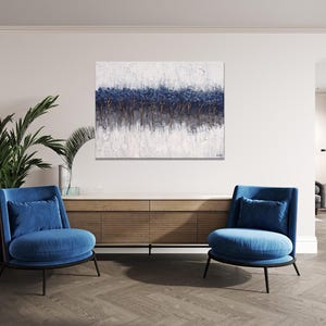 May include: Abstract art piece with a textured white background and a horizontal band of dark blue and gold tones. The artwork hangs above a wooden cabinet, flanked by two blue armchairs.