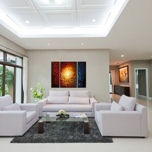 May include: A living room with white walls and a white ceiling. There are three white couches and a glass coffee table in the center of the room. A large abstract painting with red, orange, and blue colors hangs on the wall behind the couches.