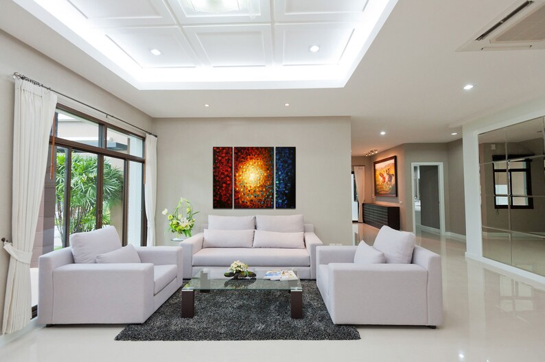 May include: A living room with white walls and a white ceiling. There are three white couches and a glass coffee table in the center of the room. A large abstract painting with red, orange, and blue colors hangs on the wall behind the couches.