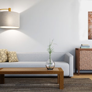 May include: A living room interior with a light gray sofa, two patterned throw pillows, a wooden coffee table, a floor lamp with a gray shade, a vase with a green plant, a wooden cabinet with a woven front, and a painting with a brown and white abstract design.