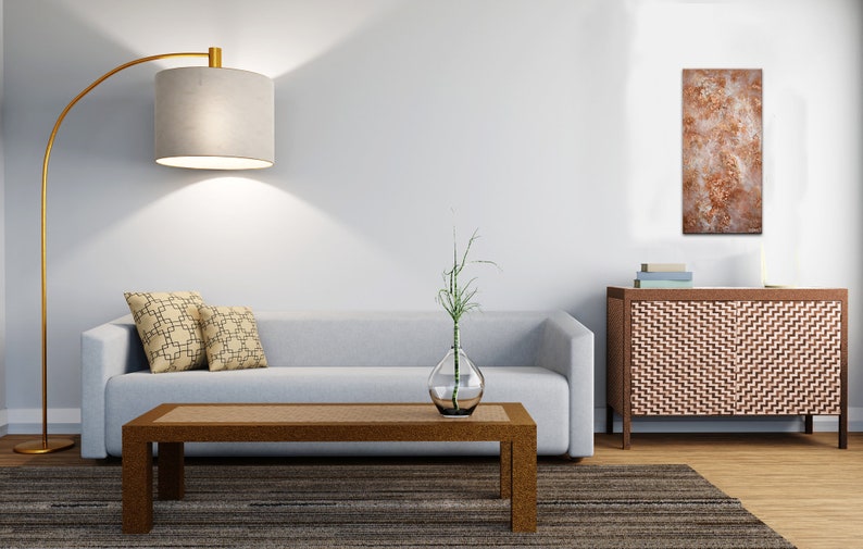 May include: A living room interior with a light gray sofa, two patterned throw pillows, a wooden coffee table, a floor lamp with a gray shade, a vase with a green plant, a wooden cabinet with a woven front, and a painting with a brown and white abstract design.