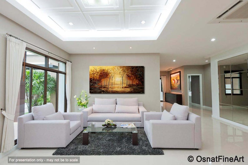 May include: A living room interior with a large abstract painting of a forest scene in shades of gold, brown, and green. The painting is hanging on a wall above a white sofa. There are two white armchairs in the room, and a coffee table with a glass top.