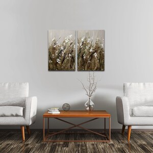 May include: Two framed paintings of a field of white flowers with brown stems and a light gray background. The paintings are hanging on a white wall above a brown coffee table and two white armchairs.