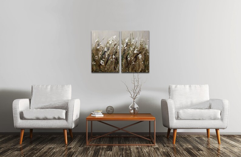 May include: Two framed paintings of a field of white flowers with brown stems and a light gray background. The paintings are hanging on a white wall above a brown coffee table and two white armchairs.