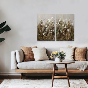 May include: A set of two abstract landscape paintings featuring a field of white flowers and brown grasses. The paintings are framed and hung on a white wall above a light gray couch with brown throw pillows.