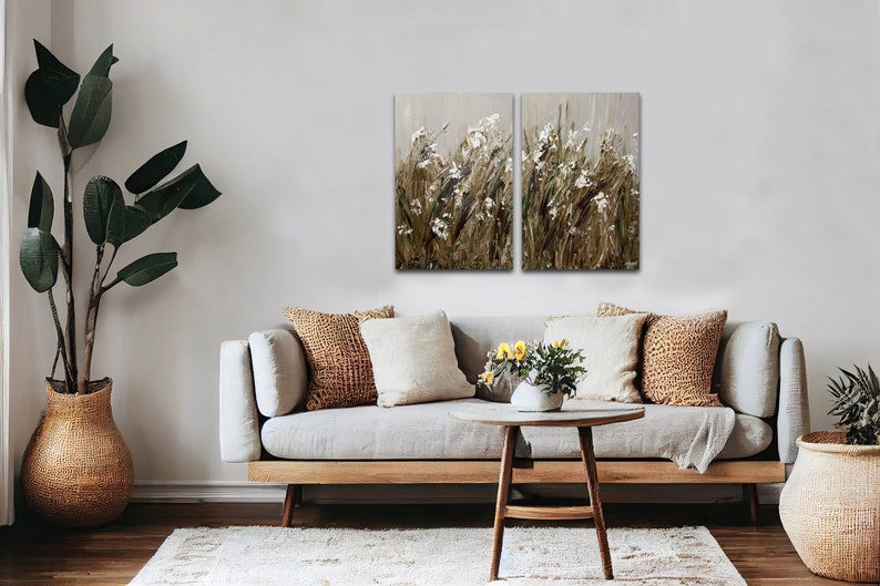 May include: A set of two abstract landscape paintings featuring a field of white flowers and brown grasses. The paintings are framed and hung on a white wall above a light gray couch with brown throw pillows.