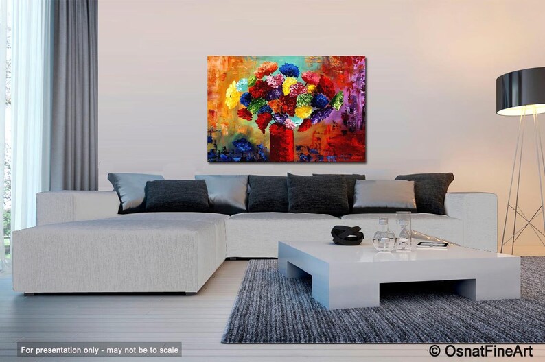 May include: An abstract painting of a bouquet of colorful flowers in a red vase. The artwork features a vibrant mix of colors, including red, yellow, blue, and purple. The painting is displayed above a white couch in a modern living room setting.