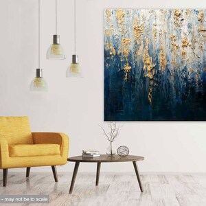 May include: Abstract textured painting with gold and navy blue tones. The artwork features vertical brushstrokes and a layered effect, creating a sense of depth. The painting is displayed in a modern living room setting with a yellow armchair and a round coffee table.