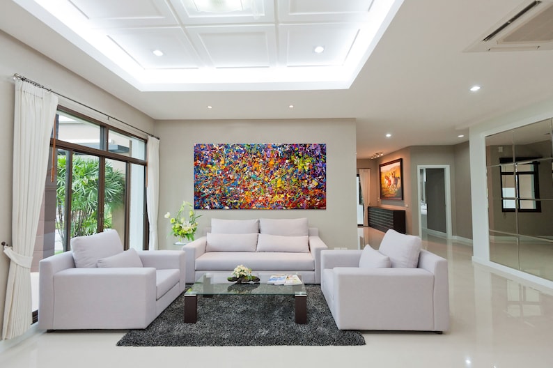 May include: A living room interior with white walls and a white ceiling. There is a large abstract painting on the wall with splashes of red, yellow, blue, green, and purple paint. The room has a white sofa, two white armchairs, a glass coffee table, and a gray rug.