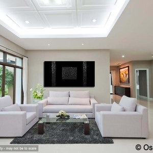 May include: A black and white abstract painting with a textured surface hangs on a wall in a modern living room. The painting is framed and is centered above a white sofa. The room has white walls, a white ceiling, and a large window with a view of green trees.