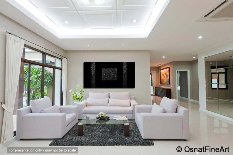 May include: A black and white abstract painting with a textured surface hangs on a wall in a modern living room. The painting is framed and is centered above a white sofa. The room has white walls, a white ceiling, and a large window with a view of green trees.