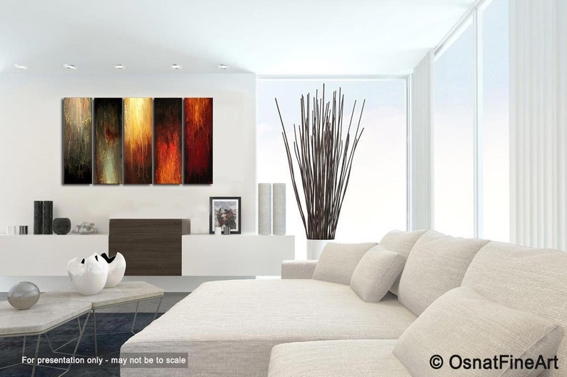 May include: Modern living room with abstract art. Five vertical panels of artwork in warm tones of gold, orange, and red hang above a white cabinet. A large white sofa with pillows and a tall plant in a white pot complete the scene.