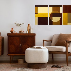 May include: A living room interior with a brown leather sofa, a white ottoman, a wooden side table, a brown armchair, and a black metal side table with a vintage rotary phone. A three-piece abstract art print with geometric shapes in shades of brown, yellow, and gold hangs on the wall.