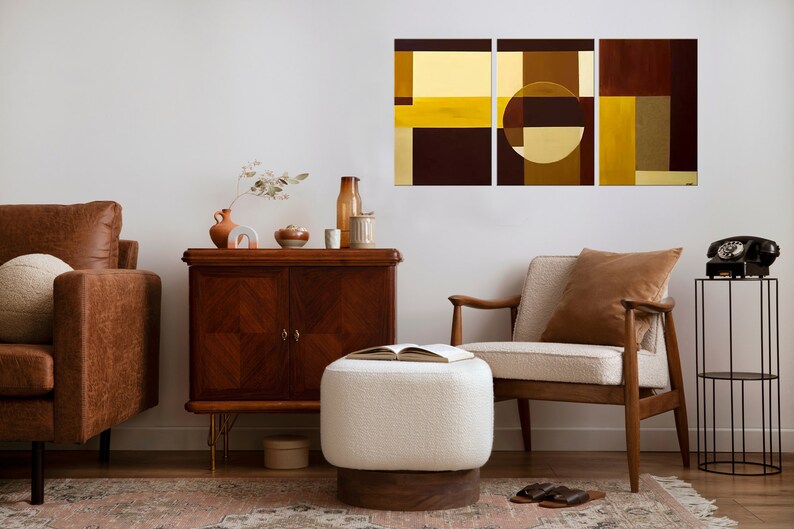 May include: A living room interior with a brown leather sofa, a white ottoman, a wooden side table, a brown armchair, and a black metal side table with a vintage rotary phone. A three-piece abstract art print with geometric shapes in shades of brown, yellow, and gold hangs on the wall.