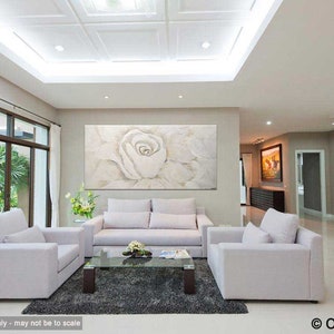 May include: A white, textured, abstract painting of a large, white rose with a light grey background. The painting is hanging on a wall in a living room with white furniture and a large window.