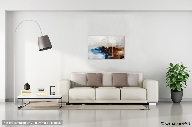 May include: Abstract painting with brown, blue, white, and gray colors. The painting is hanging on a white wall above a white couch with brown pillows.
