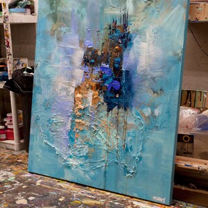 May include: Abstract painting on canvas with blue, white, and gold paint. The painting has a textured surface and is leaning against a wall.