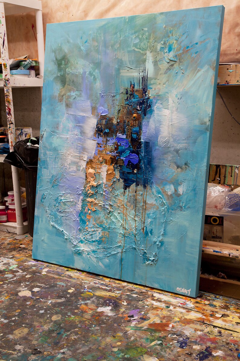 May include: Abstract painting on canvas with blue, white, and gold paint. The painting has a textured surface and is leaning against a wall.