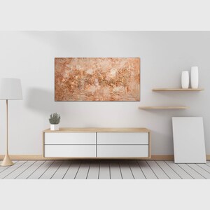 May include: An abstract painting with a textured surface in shades of brown, beige, and gold hangs on a white wall. The artwork is displayed above a modern, white cabinet with wooden accents. The room features a floor lamp and decorative shelves.