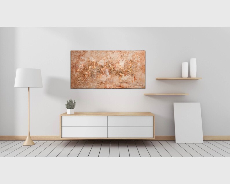 May include: An abstract painting with a textured surface in shades of brown, beige, and gold hangs on a white wall. The artwork is displayed above a modern, white cabinet with wooden accents. The room features a floor lamp and decorative shelves.