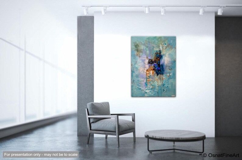 May include: Abstract painting with blue, white, and gold hues. The painting is displayed on a white wall in a modern interior setting with a gray armchair and a round coffee table.