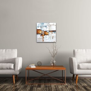 May include: Abstract painting with white, blue, and brown paint strokes. The painting is framed and hanging on a white wall above a brown coffee table.