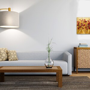 May include: A living room interior with a light gray sofa, two patterned throw pillows, a wooden coffee table, a floor lamp, a vase with a green plant, and a painting of red flowers in a field of yellow grass hanging on the wall.