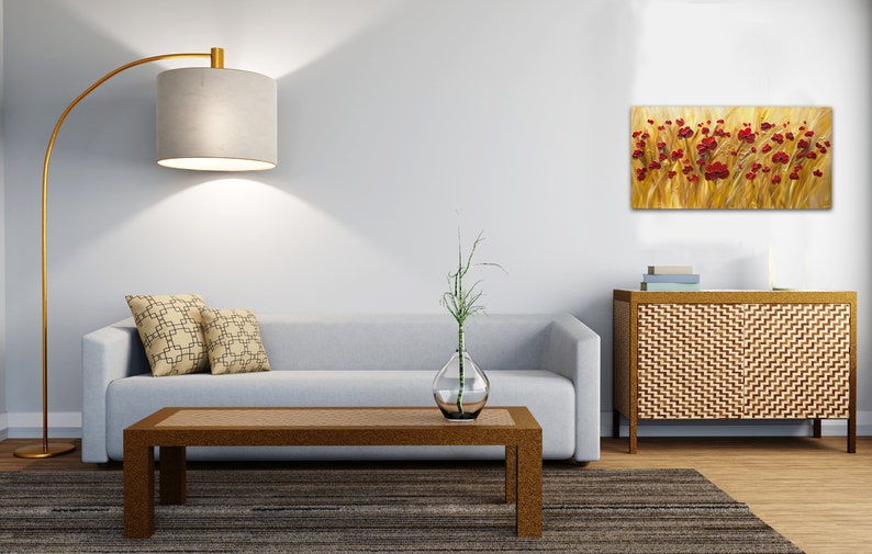 May include: A living room interior with a light gray sofa, two patterned throw pillows, a wooden coffee table, a floor lamp, a vase with a green plant, and a painting of red flowers in a field of yellow grass hanging on the wall.