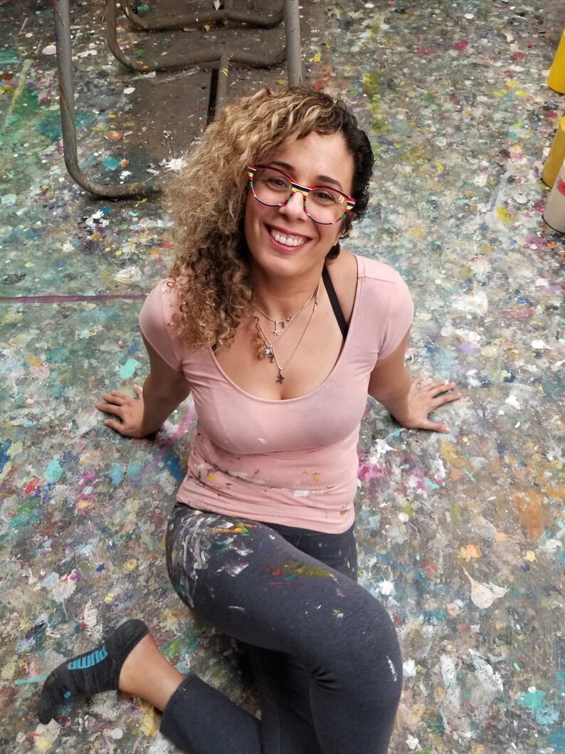 May include: A woman with curly blonde hair wearing pink and grey clothing sits on a floor covered in paint splatters. She is wearing glasses with a colorful frame and smiling at the camera.