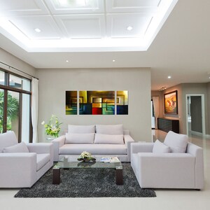 May include: A living room with white walls and a white ceiling. There is a white sofa with three cushions and a glass coffee table in front of it. There are two white armchairs on either side of the sofa. There is a large abstract painting on the wall behind the sofa. The painting is made up of squares of different colors, including yellow, red, blue, and green.