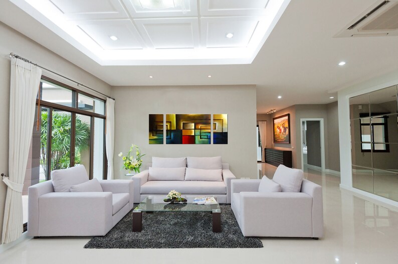 May include: A living room with white walls and a white ceiling. There is a white sofa with three cushions and a glass coffee table in front of it. There are two white armchairs on either side of the sofa. There is a large abstract painting on the wall behind the sofa. The painting is made up of squares of different colors, including yellow, red, blue, and green.