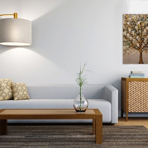 May include: A living room interior with a light gray sofa, two patterned throw pillows, a wooden coffee table, a floor lamp with a gray shade, a vase with a green plant, a wooden cabinet with a woven front, and a painting of a gold tree on the wall.