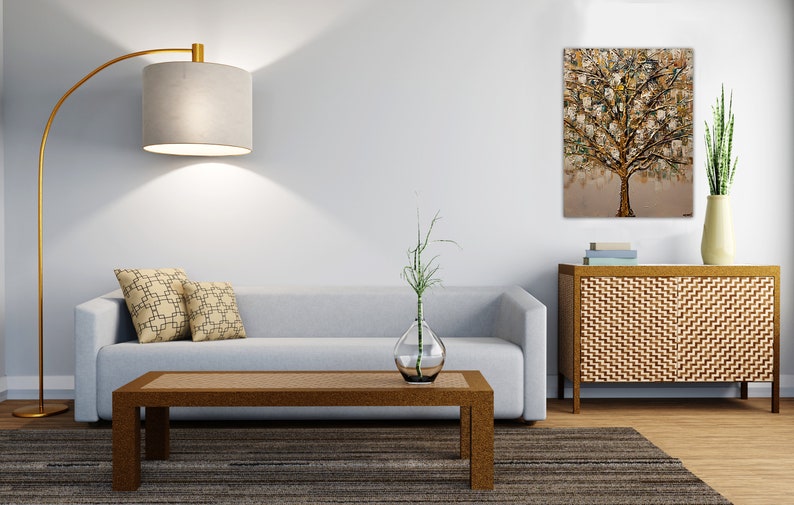 May include: A living room interior with a light gray sofa, two patterned throw pillows, a wooden coffee table, a floor lamp with a gray shade, a vase with a green plant, a wooden cabinet with a woven front, and a painting of a gold tree on the wall.