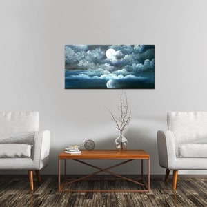 May include: A painting of a full moon shining through dark clouds over a blue ocean. The painting is framed and hanging on a white wall above a wooden coffee table with two white armchairs on either side.