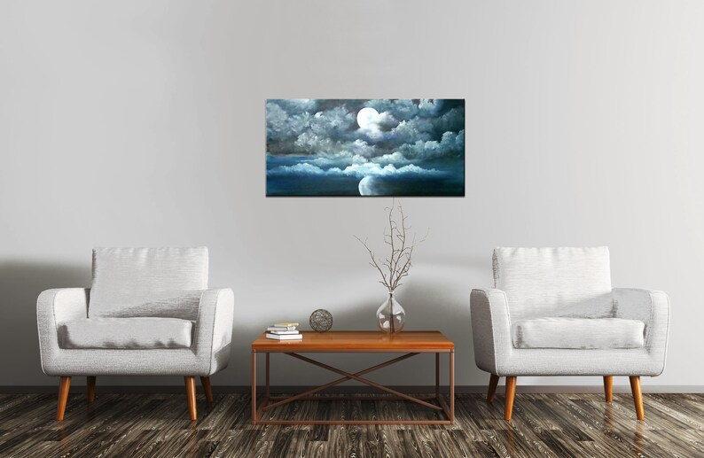 May include: A painting of a full moon shining through dark clouds over a blue ocean. The painting is framed and hanging on a white wall above a wooden coffee table with two white armchairs on either side.