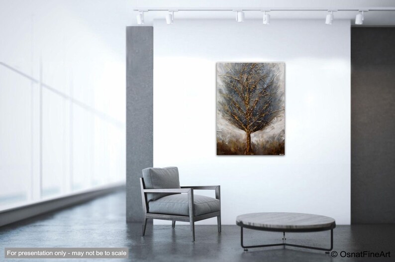 May include: A textured abstract painting of a tree with gold and silver accents. The tree is set against a background of muted gray and brown tones. The painting is displayed in a modern interior setting with a gray armchair and a round coffee table.