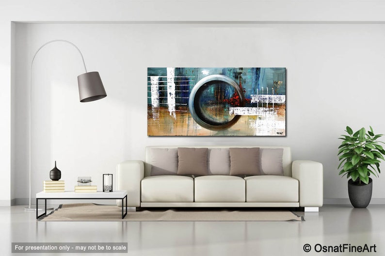 May include: Abstract painting with a large, circular, metallic gray shape in the center. The painting is on a white wall above a white couch with brown throw pillows. The painting has a blue, gray, white, and brown color scheme.