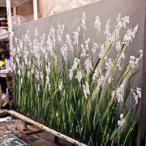May include: A large abstract painting of white flowers with green stems on a gray background. The painting is on a canvas and is leaning against a wooden easel.