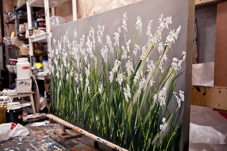May include: A large abstract painting of white flowers with green stems on a gray background. The painting is on a canvas and is leaning against a wooden easel.