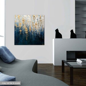 May include: Abstract painting with gold leaf accents over a dark blue and white background. The artwork is displayed in a modern living room setting, with a gray sofa and black cat figurines on a white mantel. The painting is signed in the bottom right corner.